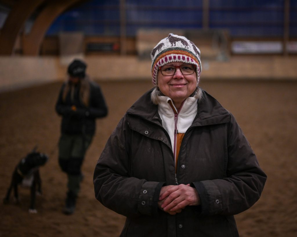 Maritha i mössa och jacka med hund i bakgrunden